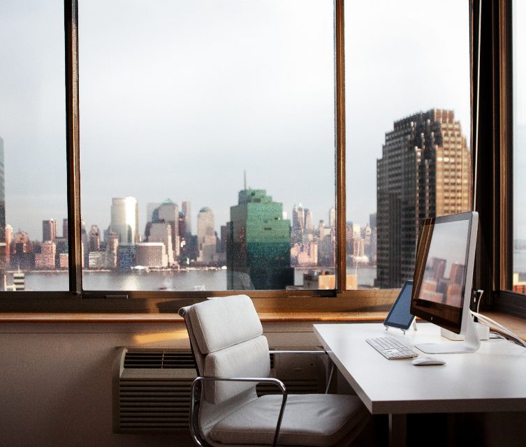 A desk with a monitor beside a large window looking out over a city